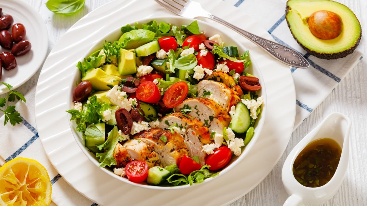 Mediterranean Rotisserie Chicken Salad with avocado, lettuce, kalamata olives, cucumber, tomato and crumbled feta cheese in white bowl on white wood table with ingredients