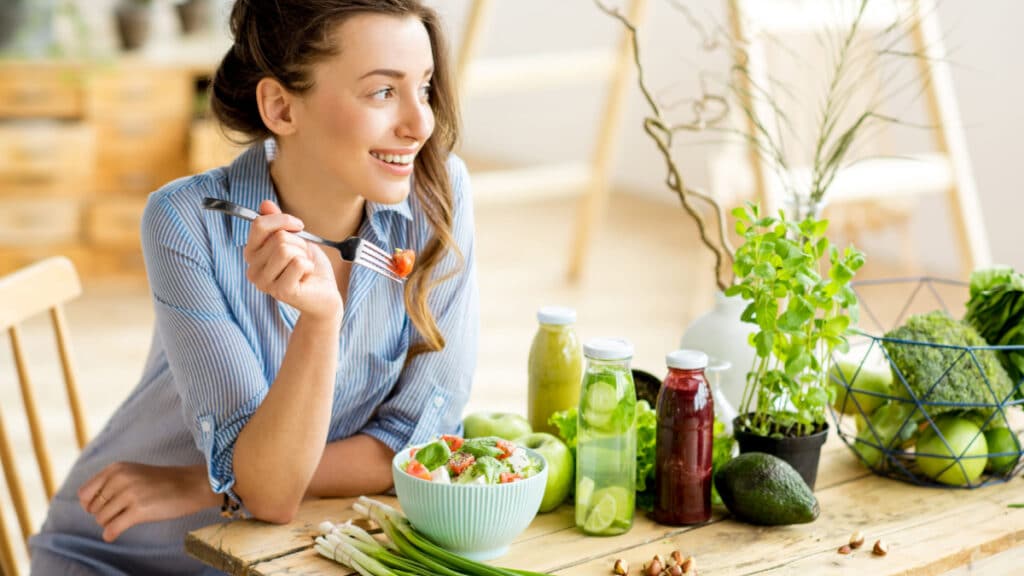 Young and happy woman eating healthy salad sitting on the table with green fresh ingredients indoors