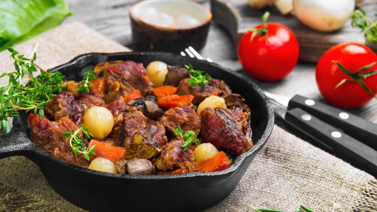 Cooking Stew meat in Burgundy (Beef Bourguignon) in cast iron frying pan with carrots, onions. Spices for Beef Bourguignon thyme, cherry tomatoes, mushrooms champignons.